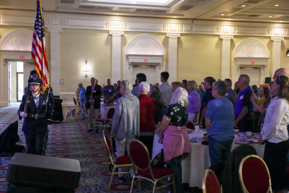 A formal indoor event is taking place in an elegant ballroom or banquet hall, where a color guard member in military dress uniform is presenting the American flag to a standing audience. The crowd of approximately 30-50 attendees is standing at attention, likely during the Pledge of Allegiance or National Anthem. The setting features ornate architectural details, round banquet tables, and red-carpeted flooring consistent with a hotel or conference center.