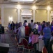 A formal indoor event is taking place in an elegant ballroom or banquet hall, where a color guard member in military dress uniform is presenting the American flag to a standing audience. The crowd of approximately 30-50 attendees is standing at attention, likely during the Pledge of Allegiance or National Anthem. The setting features ornate architectural details, round banquet tables, and red-carpeted flooring consistent with a hotel or conference center.