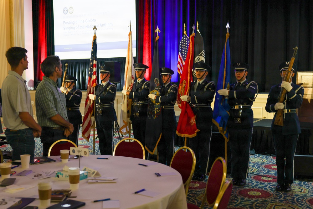 A military color guard ceremony is taking place in a banquet hall or conference room setting. Several uniformed service members, appearing to be U.S. Air Force personnel, are performing a 'Posting of the Colors' ceremony as indicated by the projection screen in the background. Two civilians stand at attention observing the ceremony, while round banquet tables with place settings are visible in the foreground.