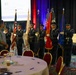 A military color guard ceremony is taking place in a banquet hall or conference room setting. Several uniformed service members, appearing to be U.S. Air Force personnel, are performing a 'Posting of the Colors' ceremony as indicated by the projection screen in the background. Two civilians stand at attention observing the ceremony, while round banquet tables with place settings are visible in the foreground.