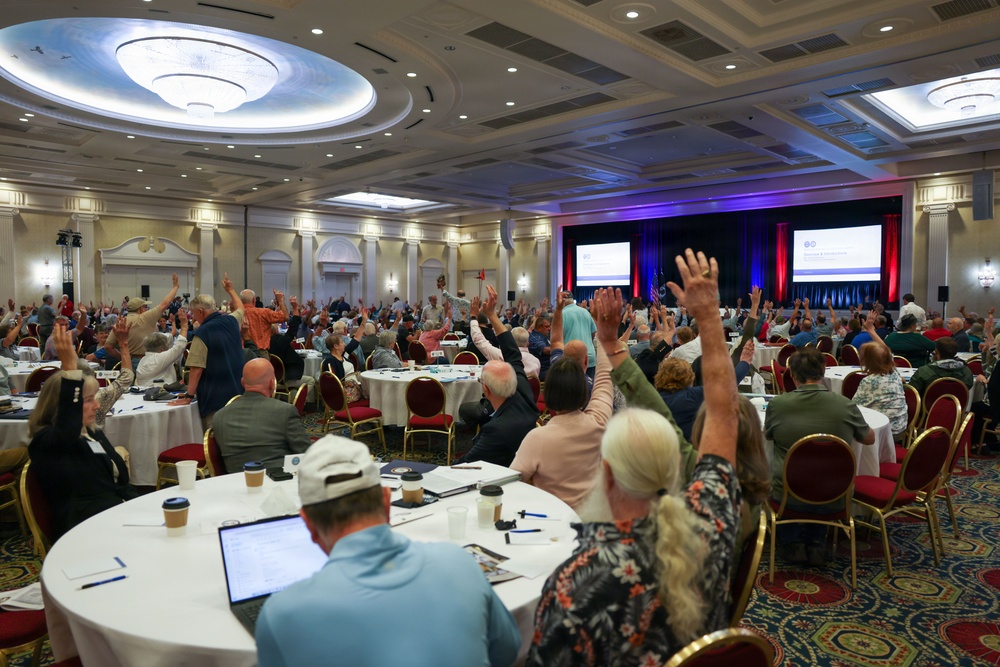 A large formal conference or convention is taking place in an upscale hotel ballroom, with hundreds of attendees seated at round tables raising their hands, likely in response to a vote or poll. The room features elegant decor including coffered ceilings with chandeliers, and a stage at the front with large projection screens displaying presentation content. The event appears to be an organized assembly or annual meeting of a professional or civic organization.