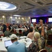 A large formal conference or convention is taking place in an upscale hotel ballroom, with hundreds of attendees seated at round tables raising their hands, likely in response to a vote or poll. The room features elegant decor including coffered ceilings with chandeliers, and a stage at the front with large projection screens displaying presentation content. The event appears to be an organized assembly or annual meeting of a professional or civic organization.