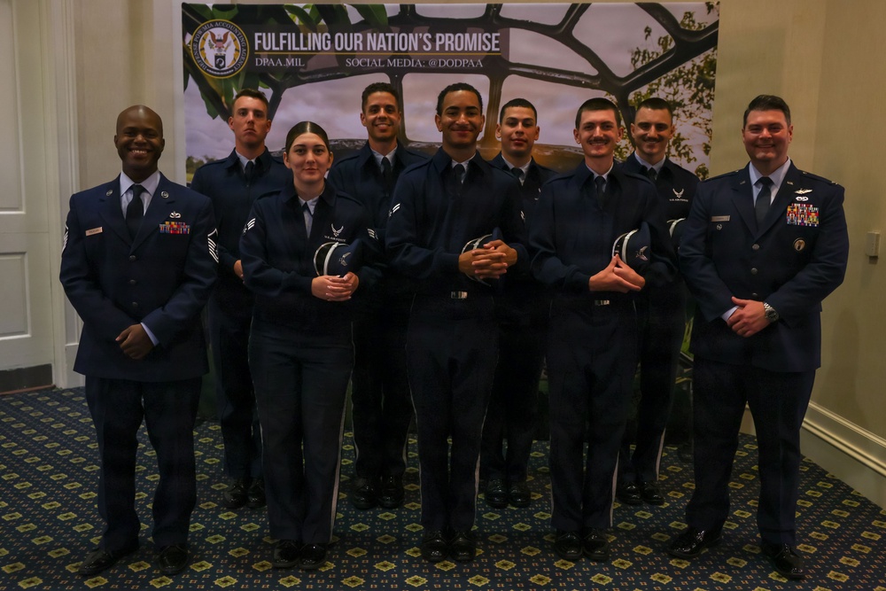 A group of nine U.S. Air Force personnel in dress blue uniforms pose for a formal group photo in front of a DPAA (Defense POW/MIA Accounting Agency) banner reading 'Fulfilling Our Nation's Promise.' Several members are holding what appear to be awards or recognition items. The setting appears to be an indoor ceremonial or awards event.