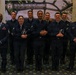 A group of nine U.S. Air Force personnel in dress blue uniforms pose for a formal group photo in front of a DPAA (Defense POW/MIA Accounting Agency) banner reading 'Fulfilling Our Nation's Promise.' Several members are holding what appear to be awards or recognition items. The setting appears to be an indoor ceremonial or awards event.