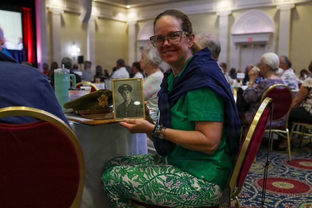 A woman wearing glasses and a green outfit sits at a banquet-style event, proudly holding a framed black-and-white portrait photograph of a military officer in uniform. On the table beside her rests what appears to be a vintage U.S. Army officer's service cap with an eagle insignia. The setting appears to be a formal gathering or commemorative event held in a large ballroom with many attendees in the background.
