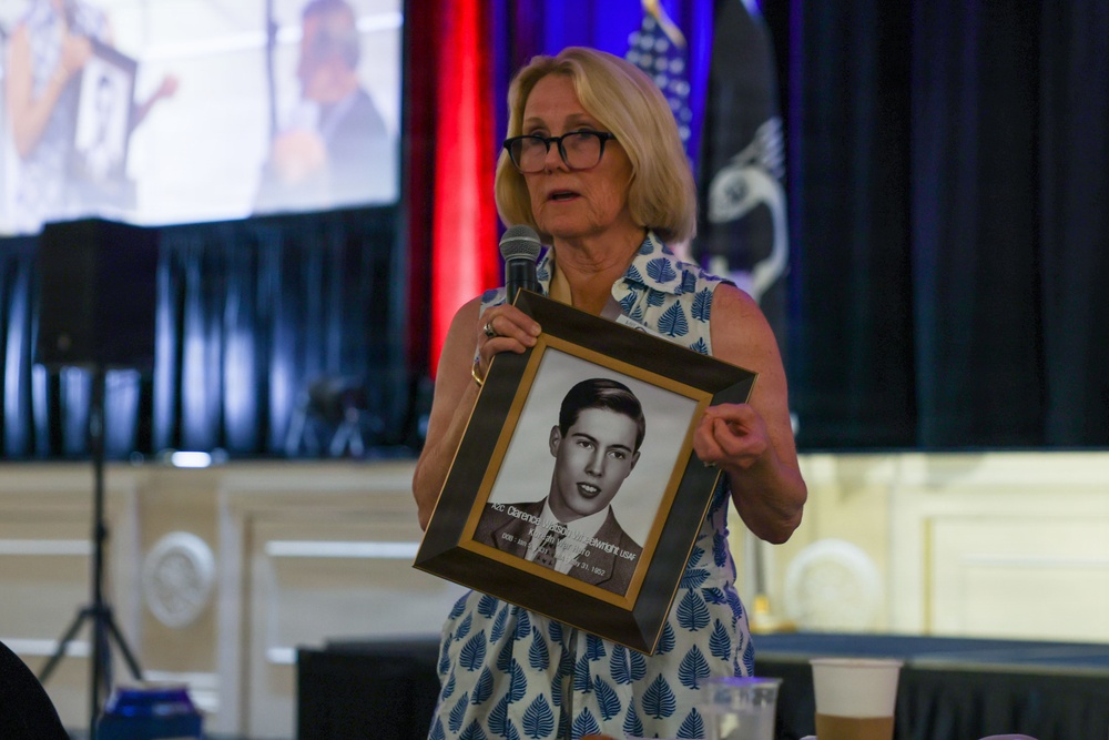 A woman with blonde hair and glasses is speaking at a formal event while holding a framed black-and-white portrait photograph of a young man. The framed photo appears to have a caption/plaque identifying a military servicemember, partially readable as 'Clarence' and 'USAF'. The setting appears to be a banquet hall or ceremonial venue with patriotic red, white, and blue decorations and flags in the background.