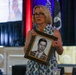 A woman with blonde hair and glasses is speaking at a formal event while holding a framed black-and-white portrait photograph of a young man. The framed photo appears to have a caption/plaque identifying a military servicemember, partially readable as 'Clarence' and 'USAF'. The setting appears to be a banquet hall or ceremonial venue with patriotic red, white, and blue decorations and flags in the background.