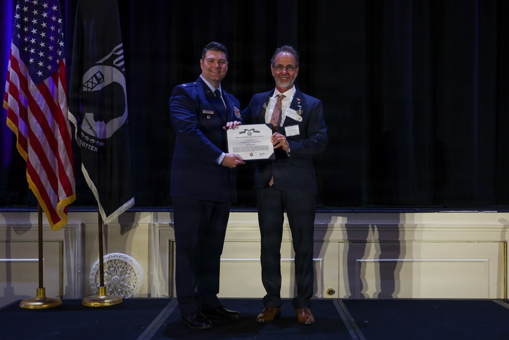 Two men stand on a stage holding a certificate or award document between them. One man is wearing a U.S. Air Force dress uniform while the other is in civilian business attire with a name badge and medal/pin. The setting appears to be a formal awards ceremony with an American flag and POW/MIA flag visible in the background.