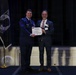 Two men stand on a stage holding a certificate or award document between them. One man is wearing a U.S. Air Force dress uniform while the other is in civilian business attire with a name badge and medal/pin. The setting appears to be a formal awards ceremony with an American flag and POW/MIA flag visible in the background.