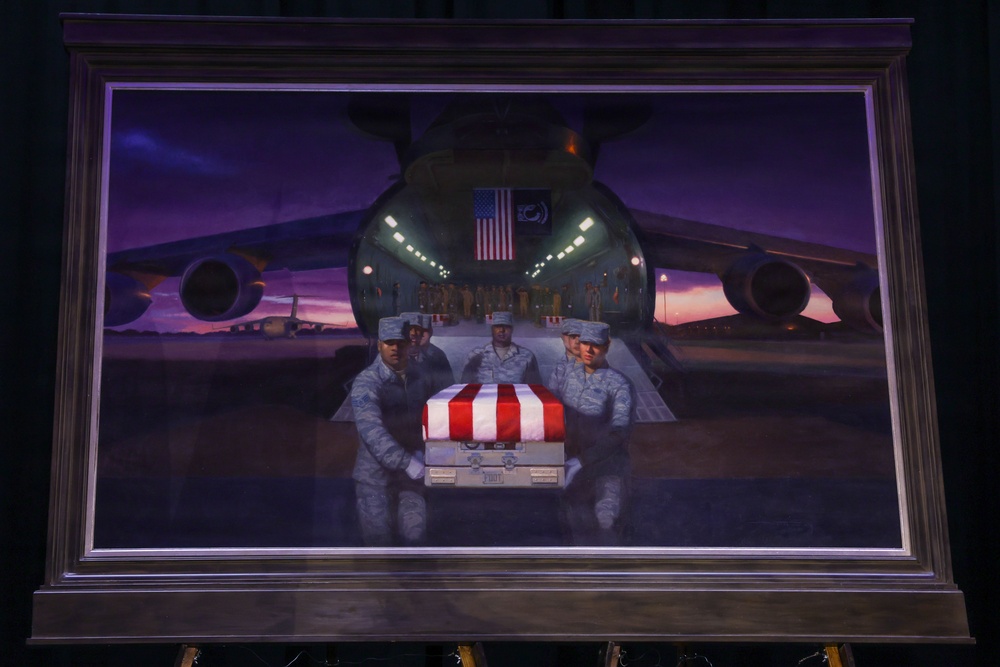 A framed painting displayed on an easel depicting a military dignified transfer ceremony at an airfield. The artwork shows uniformed service members carrying a flag-draped transfer case from the rear cargo bay of a large military transport aircraft at dusk or dawn. The painting uses dramatic purple and orange tones to convey a solemn, ceremonial atmosphere.