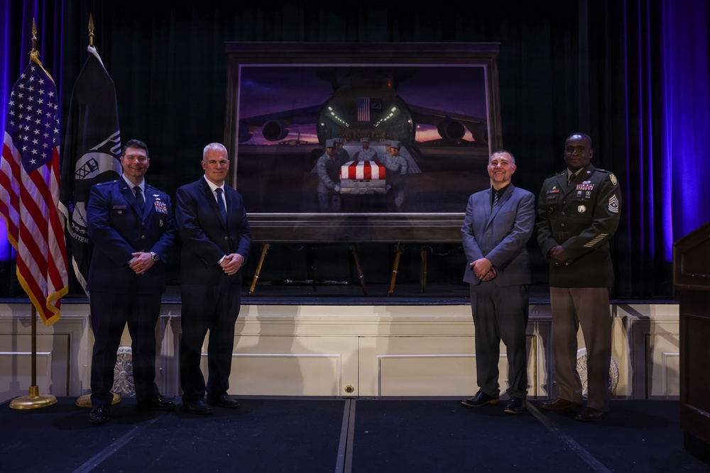 Four individuals stand in front of a large framed painting on a stage during what appears to be a formal military or commemorative ceremony. The painting depicts uniformed personnel carrying a flag-draped casket from a military aircraft, suggesting a dignified transfer or repatriation scene. Two individuals are in military uniform (U.S. Air Force and U.S. Army) and two are in civilian attire.