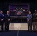 Four individuals stand in front of a large framed painting on a stage during what appears to be a formal military or commemorative ceremony. The painting depicts uniformed personnel carrying a flag-draped casket from a military aircraft, suggesting a dignified transfer or repatriation scene. Two individuals are in military uniform (U.S. Air Force and U.S. Army) and two are in civilian attire.