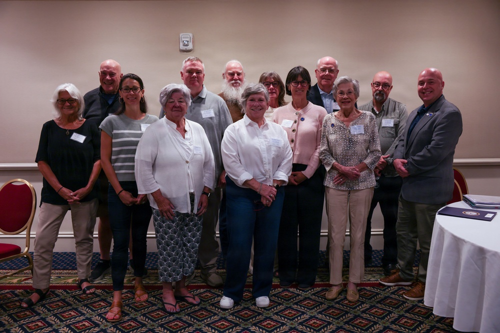 A group of approximately 13 adults posing together for a formal group photograph in what appears to be a banquet or conference room. Most individuals are wearing name tags, suggesting this is an organized event such as a meeting, awards ceremony, or organizational gathering. The group consists of mixed genders and appears to be predominantly older adults.