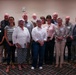 A group of approximately 13 adults posing together for a formal group photograph in what appears to be a banquet or conference room. Most individuals are wearing name tags, suggesting this is an organized event such as a meeting, awards ceremony, or organizational gathering. The group consists of mixed genders and appears to be predominantly older adults.