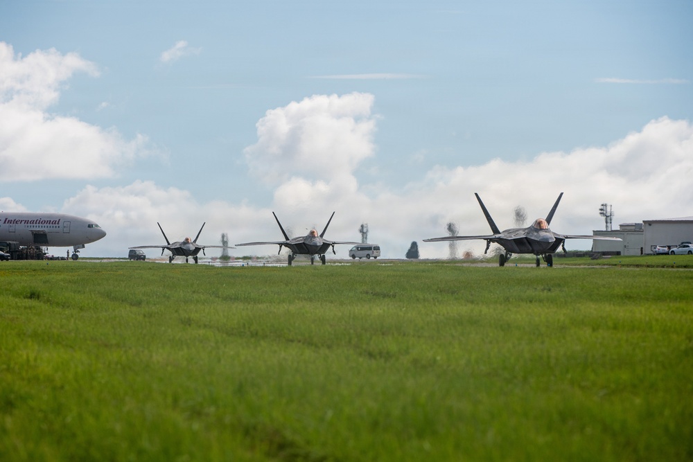 Three stealth fighter jets, consistent with F-22 Raptors, are taxiing in formation on a wet airfield taxiway. A large commercial or cargo aircraft with 'International' visible on its fuselage is parked on the left side of the frame. The scene appears to be at a joint-use or military airfield with support vehicles and infrastructure visible in the background.