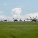 Three stealth fighter jets, consistent with F-22 Raptors, are taxiing in formation on a wet airfield taxiway. A large commercial or cargo aircraft with 'International' visible on its fuselage is parked on the left side of the frame. The scene appears to be at a joint-use or military airfield with support vehicles and infrastructure visible in the background.