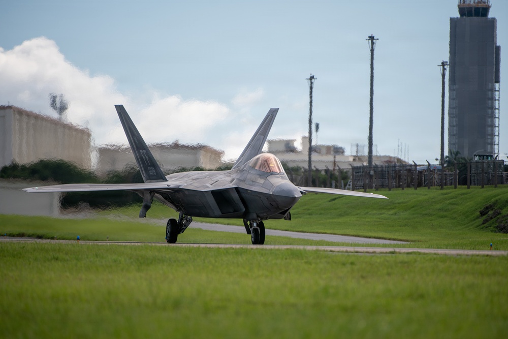 A U.S. Air Force F-22 Raptor stealth fighter jet is taxiing on a runway at what appears to be a military airfield. The aircraft is viewed from a front-facing angle, showing its distinctive twin-tail and blended wing design. An air traffic control tower and hardened aircraft shelters are visible in the background.