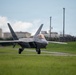A U.S. Air Force F-22 Raptor stealth fighter jet is taxiing on a runway at what appears to be a military airfield. The aircraft is viewed from a front-facing angle, showing its distinctive twin-tail and blended wing design. An air traffic control tower and hardened aircraft shelters are visible in the background.