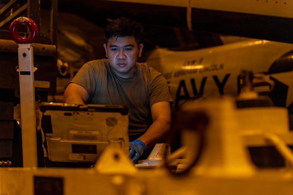 A U.S. Navy sailor in an olive/green t-shirt works in what appears to be a ship's hangar bay, likely aboard the USS Abraham Lincoln based on visible text in the background. The individual is wearing blue nitrile gloves and appears to be performing maintenance or equipment checks on military hardware. The scene is illuminated by warm amber/orange lighting typical of nighttime hangar deck operations.