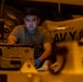 A U.S. Navy sailor in an olive/green t-shirt works in what appears to be a ship's hangar bay, likely aboard the USS Abraham Lincoln based on visible text in the background. The individual is wearing blue nitrile gloves and appears to be performing maintenance or equipment checks on military hardware. The scene is illuminated by warm amber/orange lighting typical of nighttime hangar deck operations.