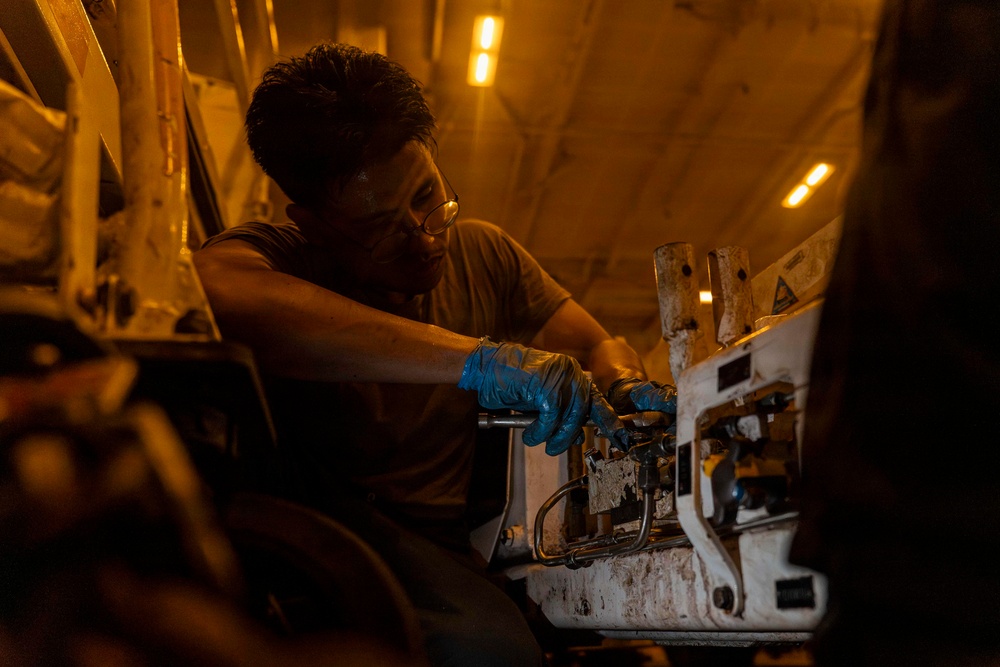 A technician wearing blue nitrile gloves and glasses is performing maintenance or repair work on a complex mechanical or hydraulic assembly in a dimly lit industrial or aircraft interior environment. The warm amber lighting from overhead fixtures casts dramatic shadows across the workspace. The worker appears focused and is using a hand tool on what appears to be a valve or hydraulic manifold system.