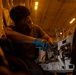 A technician wearing blue nitrile gloves and glasses is performing maintenance or repair work on a complex mechanical or hydraulic assembly in a dimly lit industrial or aircraft interior environment. The warm amber lighting from overhead fixtures casts dramatic shadows across the workspace. The worker appears focused and is using a hand tool on what appears to be a valve or hydraulic manifold system.