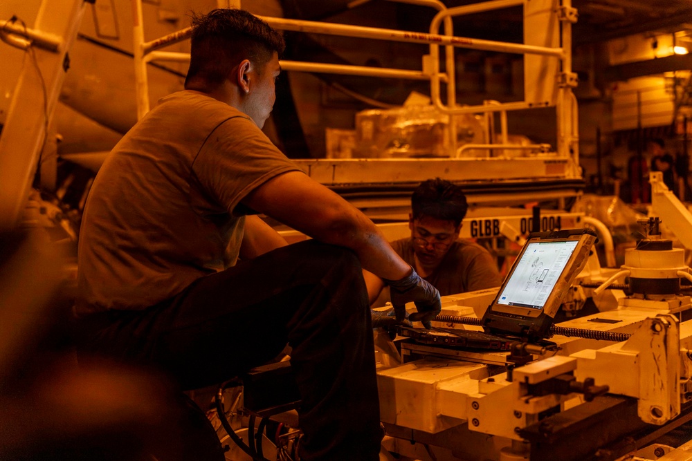 Two individuals in military-style brown t-shirts are working on heavy equipment in a dimly lit industrial or military vehicle bay. They are using a ruggedized laptop/tablet computer, likely referencing technical diagrams or schematics while performing maintenance. The environment is bathed in an orange ambient light, suggesting sodium vapor or similar industrial lighting.