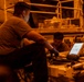 Two individuals in military-style brown t-shirts are working on heavy equipment in a dimly lit industrial or military vehicle bay. They are using a ruggedized laptop/tablet computer, likely referencing technical diagrams or schematics while performing maintenance. The environment is bathed in an orange ambient light, suggesting sodium vapor or similar industrial lighting.