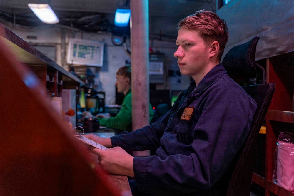 A young person in a dark navy blue uniform or coveralls sits in a chair holding a document or paper, appearing focused and attentive in what looks like an industrial or military workspace. In the background, a second individual wearing a green shirt works at a workstation with equipment and displays. The environment is dimly lit with mixed red and blue ambient lighting, suggesting a control room or operations center.
