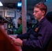 A young person in a dark navy blue uniform or coveralls sits in a chair holding a document or paper, appearing focused and attentive in what looks like an industrial or military workspace. In the background, a second individual wearing a green shirt works at a workstation with equipment and displays. The environment is dimly lit with mixed red and blue ambient lighting, suggesting a control room or operations center.
