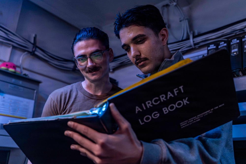 Two individuals are examining an Aircraft Log Book together in a dimly lit, industrial-looking space. The person on the right is holding the black logbook, which appears to be an official United States Navy document, while the person on the left observes. The setting and document suggest a military aviation maintenance or administrative context.