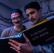 Two individuals are examining an Aircraft Log Book together in a dimly lit, industrial-looking space. The person on the right is holding the black logbook, which appears to be an official United States Navy document, while the person on the left observes. The setting and document suggest a military aviation maintenance or administrative context.