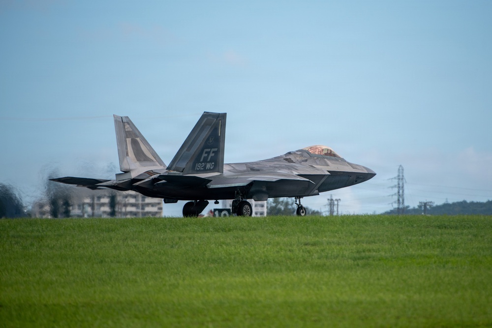 An F-22 Raptor stealth fighter jet is taxiing on a runway, displaying tail markings 'FF' and '192 WG', indicating it belongs to the 192nd Wing of the Virginia Air National Guard based at Langley Air Force Base. The aircraft is photographed from a low angle on a grass-lined airfield under overcast skies. Heat distortion or exhaust is visible behind the aircraft's engines.