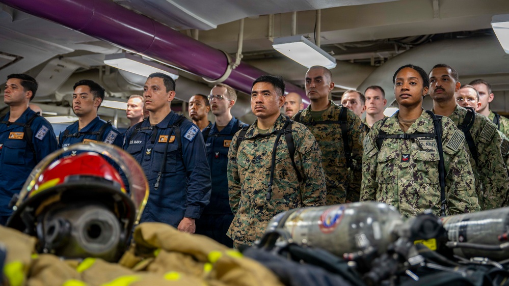 A group of U.S. military personnel from multiple branches, including the U.S. Navy and U.S. Marine Corps, are standing at attention in what appears to be the interior of a naval vessel. Firefighting and damage control equipment, including SCBA tanks, a firefighting helmet, and turnout gear, are visible in the foreground. The setting suggests a damage control or firefighting training briefing aboard a ship.