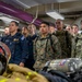 A group of U.S. military personnel from multiple branches, including the U.S. Navy and U.S. Marine Corps, are standing at attention in what appears to be the interior of a naval vessel. Firefighting and damage control equipment, including SCBA tanks, a firefighting helmet, and turnout gear, are visible in the foreground. The setting suggests a damage control or firefighting training briefing aboard a ship.