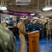 A senior military officer in tan/khaki service uniform is addressing a formation of sailors and Marines from a wooden podium aboard what appears to be a U.S. Navy vessel. The audience is standing at ease in a large interior compartment, likely a mess deck or hangar bay, wearing a mix of Navy working uniforms and Marine Corps combat utilities. The setting and attire suggest a formal military ceremony or all-hands briefing.