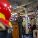 A military officer in a flight suit delivers remarks at a wooden podium aboard what appears to be a naval vessel, with an American flag displayed behind him. Two other uniformed naval personnel are present — one standing and one seated in a red chair. In the foreground, a red and yellow firefighting/damage control helmet is visible, suggesting a shipboard ceremony or briefing.