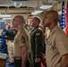 A military ceremony is taking place in what appears to be a ship's recreation or dining facility, with several uniformed personnel standing at attention. Multiple branches are represented, including U.S. Marines and what appears to be a naval aviator in a green flight suit. An American flag and a U.S. Marine Corps flag are visible in the background.
