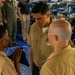 Three U.S. Navy personnel in tan/khaki uniforms are engaged in what appears to be a formal military ceremony, with one sailor holding a folded flag. A senior enlisted sailor wearing ribbons and a warfare device stands centrally, overseeing the interaction. The event takes place in an indoor hangar or bay environment with other military personnel observing in the background.