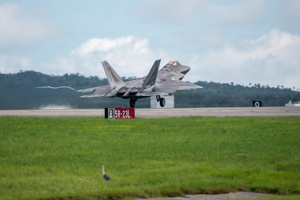 A military stealth fighter jet, consistent with an F-22 Raptor, is captured during takeoff or landing on a runway. The aircraft displays a gray camouflage paint scheme with its landing gear still deployed. A runway taxiway sign reading 'B 5R-23L' is visible in the foreground, with tropical vegetation visible in the background.
