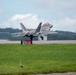 A military stealth fighter jet, consistent with an F-22 Raptor, is captured during takeoff or landing on a runway. The aircraft displays a gray camouflage paint scheme with its landing gear still deployed. A runway taxiway sign reading 'B 5R-23L' is visible in the foreground, with tropical vegetation visible in the background.