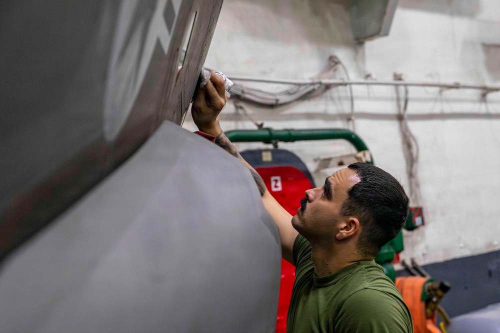 A young man in an olive green military-style t-shirt is performing maintenance work on what appears to be a military aircraft, likely in a hangar bay aboard a naval vessel. He is reaching upward to work on a panel or seam on the aircraft's fuselage, holding what appears to be a tool or cloth. The background reveals industrial infrastructure including pipes, cables, and equipment typical of a ship's hangar deck.