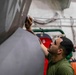 A young man in an olive green military-style t-shirt is performing maintenance work on what appears to be a military aircraft, likely in a hangar bay aboard a naval vessel. He is reaching upward to work on a panel or seam on the aircraft's fuselage, holding what appears to be a tool or cloth. The background reveals industrial infrastructure including pipes, cables, and equipment typical of a ship's hangar deck.