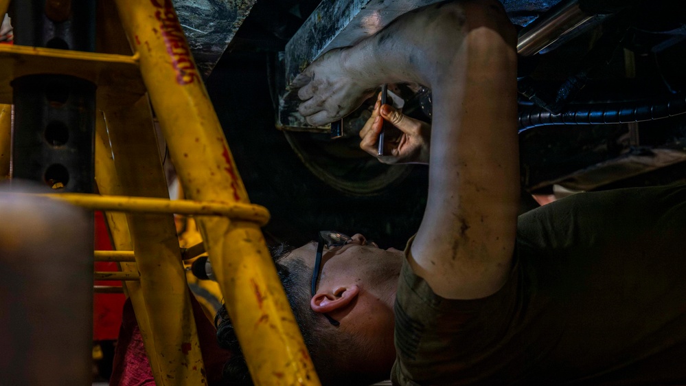 A mechanic is lying on their back underneath a vehicle, performing maintenance or repair work. They are wearing safety glasses and working with a tool in a dimly lit garage or workshop environment. A yellow safety stand or jack stand is visible nearby, supporting the vehicle.