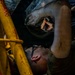 A mechanic is lying on their back underneath a vehicle, performing maintenance or repair work. They are wearing safety glasses and working with a tool in a dimly lit garage or workshop environment. A yellow safety stand or jack stand is visible nearby, supporting the vehicle.