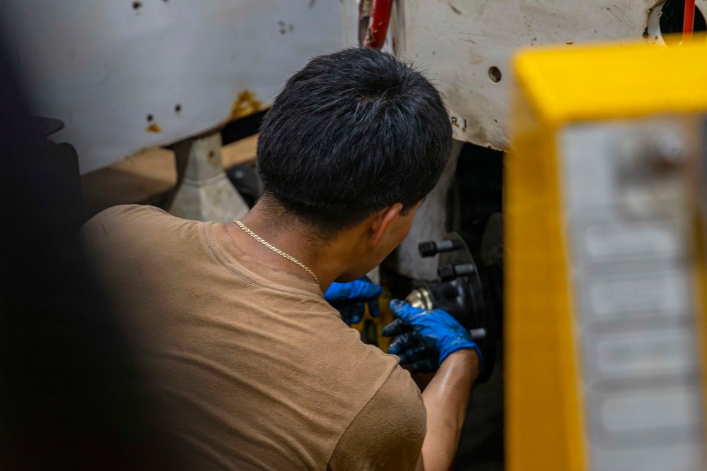 A mechanic is working on a vehicle's brake or suspension component, viewed from behind. He is wearing blue nitrile gloves and a tan t-shirt with a gold chain necklace visible. The work appears to be taking place in an automotive repair shop or garage setting.