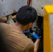 A mechanic is working on a vehicle's brake or suspension component, viewed from behind. He is wearing blue nitrile gloves and a tan t-shirt with a gold chain necklace visible. The work appears to be taking place in an automotive repair shop or garage setting.