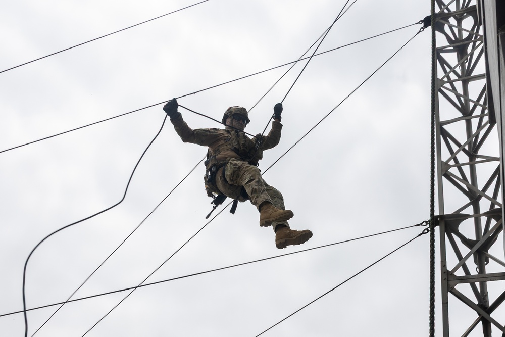 A soldier in full military camouflage uniform and helmet is suspended mid-air on ropes, appearing to conduct a rappelling or zip-line training exercise. The individual is attached to multiple lines extending from a large metal tower structure visible on the right side of the frame. The overcast sky provides a neutral background, emphasizing the elevated position of the subject.