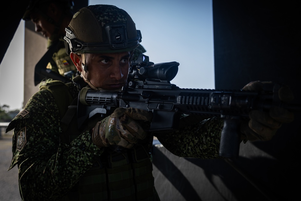 A soldier in digital camouflage uniform and tactical helmet is aiming a rifle with an optical scope attachment from a covered position. The image appears to depict a military training exercise or tactical operation, with a second soldier partially visible in the background. The dramatic lighting and composition suggest a professional military photography context.