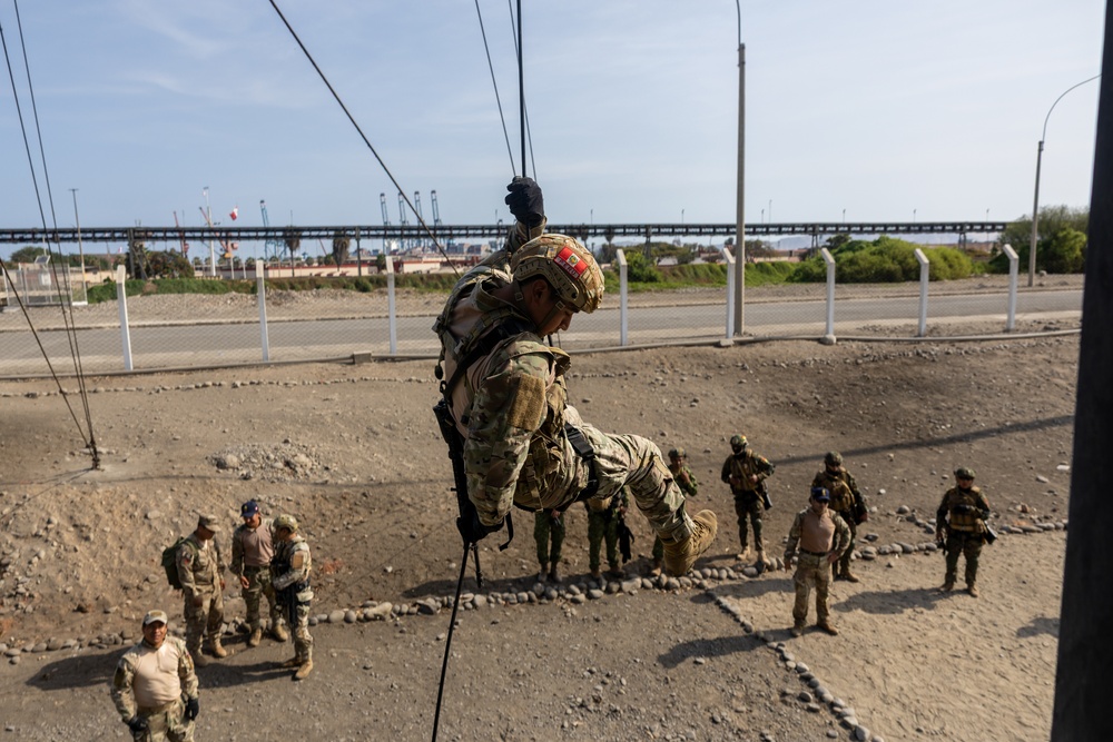 A military or special operations training exercise is underway, with one soldier rappelling or performing a rope descent from an elevated structure while carrying full combat gear. Several other uniformed personnel observe from the ground below. The setting appears to be a coastal or port-adjacent training area with industrial infrastructure visible in the background.